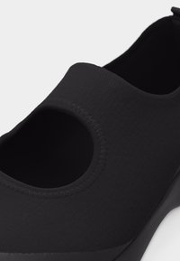 Μαύρο παπούτσι χωρίς κορδόνια με στρογγυλεμένη εγκοπή κοντά στο πάνω μέρος και ραμμένα άκρα, που δείχνει απαλό υφασμάτινο υφή.