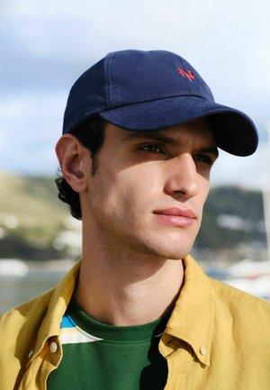 Junger Mann mit marineblauer Kappe, grünem Hemd und gelber Jacke, schaut seitlich mit unscharfem Hintergrund im Freien.