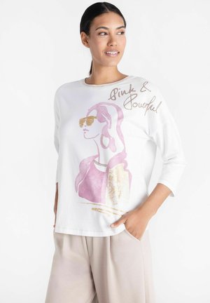 Vrouw draagt wit shirt met lange mouwen met roze en gouden afbeelding van een vrouw met zonnebril en de woorden "Pink & Powerful".