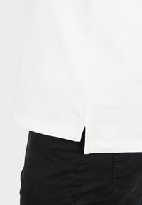 Hvid T-shirt med lige skåret kant og sideslids, lavet af struktureret stof, parret med sorte strukturerede bukser.