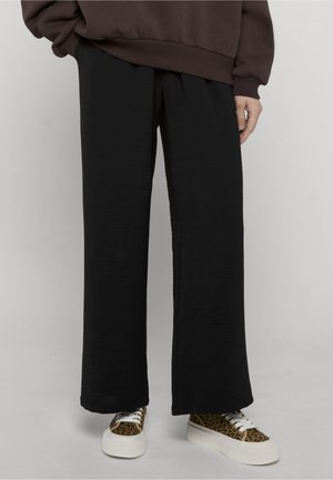 Henkilö, jolla on päällään väljät mustat housut ja leopardikuvioiset lenkkarit, joissa on paksut valkoiset pohjat, seisoo yksivärisen taustan edessä.