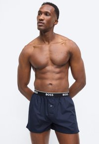 Μυώδης άνδρας χωρίς πουκάμισο που φοράει σκούρο μπλε μποξεράκια με μαύρη ζώνη μέσης που γράφει "BOSS", στέκεται με τα χέρια πίσω από την πλάτη.