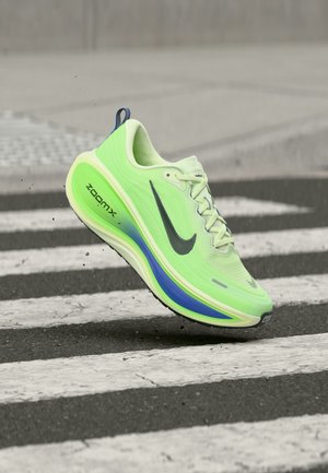 Chaussure de course Nike ZoomX vert clair en l'air au-dessus d'un passage piéton noir et blanc sur l'asphalte.