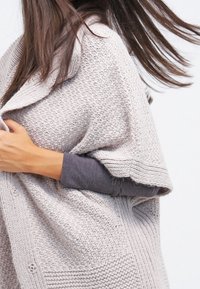 Poncho tricoté beige clair avec un motif texturé, un large col et des boutons sur les côtés. Tissu doux tombant sur les épaules.