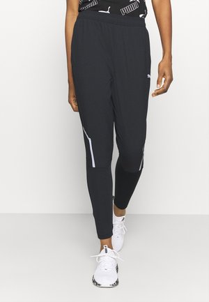Person trägt schwarze Puma-Sporthose mit reflektierenden Streifen und weiße Puma-Sneakers, steht vor einem einfarbig hellen Hintergrund.