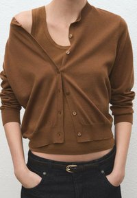 Cárdigan marrón de punto con cuello redondo, con cierres de botones y mangas largas, superpuesto a una camiseta sin mangas marrón. Tejido texturizado.