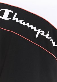 Svart och vit t-shirt med röda accenter. Har en djärv "Champion"-logotyp i vitt, kontrasterande sömmar och en sportig, modern design.