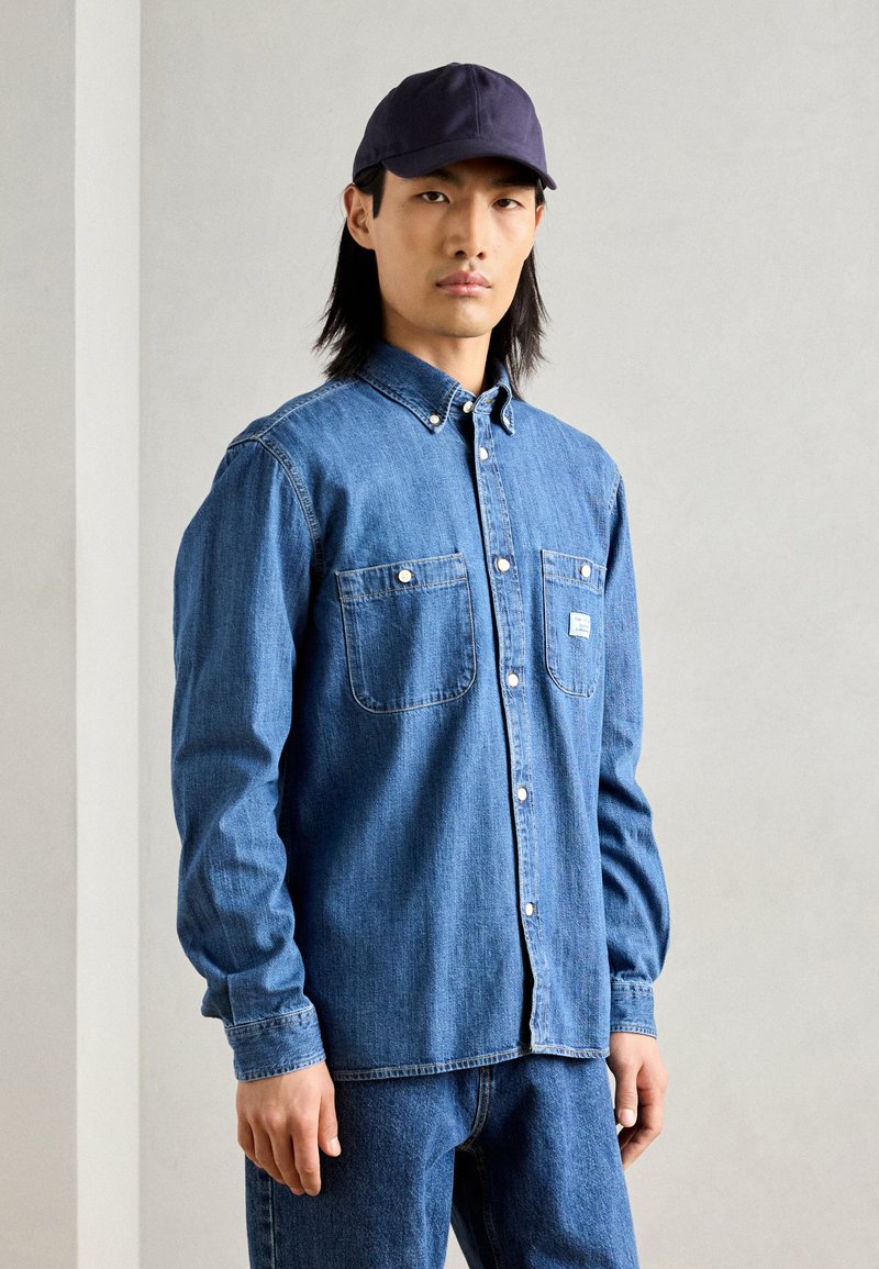 Denim shirt with a button-down collar, two chest pockets, and a slim fit, paired with dark blue jeans and a navy cap.