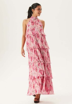 Donna sorridente in piedi, che indossa un abito lungo rosa senza maniche a balze floreali con pieghe e tacchi con plateau.