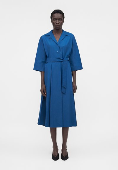 Femme portant une robe bleue ceinturée mi-longue aux manches larges et des escarpins noirs à bouts pointus, debout devant un fond blanc uni.