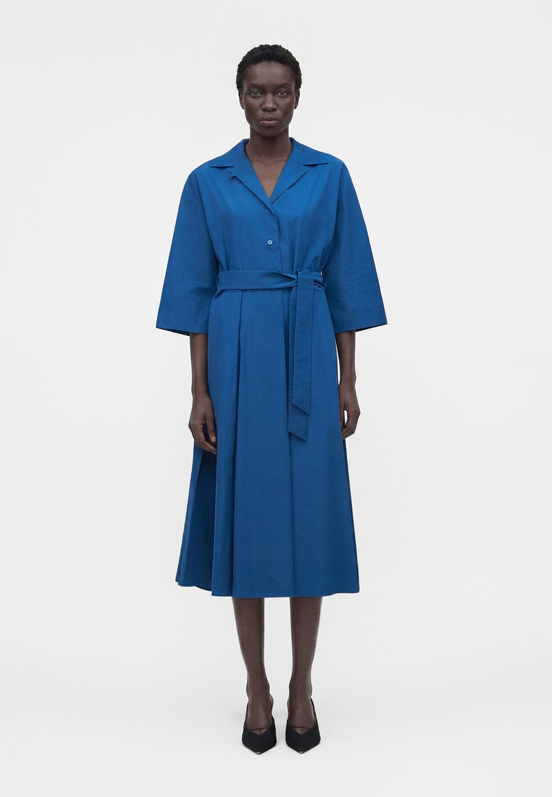 Γυναίκα που φορά φορέμα μπλε μήκους μέχρι το γόνατο με ζώνη και φαρδιά μανίκια και μαύρες μυτερές γόβες, στέκεται μπροστά σε απλό λευκό φόντο.