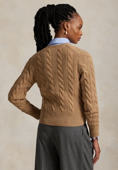 Femeie cu părul împletit, purtând un pulover bej cu model ciorchine peste o cămașă albastră cu guler și pantaloni gri, cu fața în lateral și mâna pe șold.