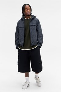 Jacket azul marinho texturizado com fecho, com dois bolsos frontais, sobre uma camisa verde-oliva, combinado com calções de carga pretos e ténis brancos.