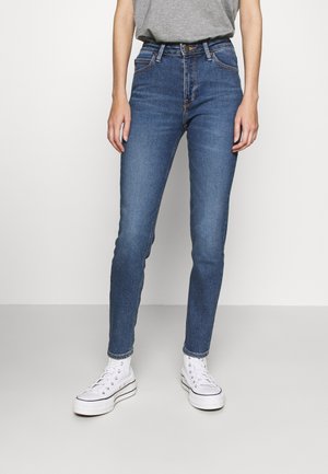 Personne portant un jean skinny bleu taille haute et des baskets blanches montantes, debout devant un fond clair uni.