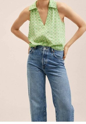 Ærmeløs lysegrøn mønstret bluse stoppet ned i høj-taljede blå denimjeans, hænderne hvilende på hofterne.