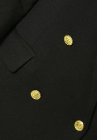 Juoda dvigubo priekinio užsegimo audinio striukė su trimis auksinėmis reljefinėmis sagomis ir audinio sklende virš viršutinės sagos.