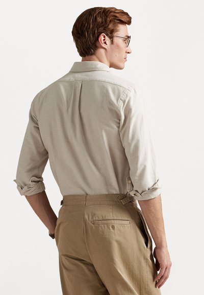 Light beige button-up shirt with rolled sleeves, featuring a back pleat. Paired with khaki trousers, showcasing belt loops and a back pocket.