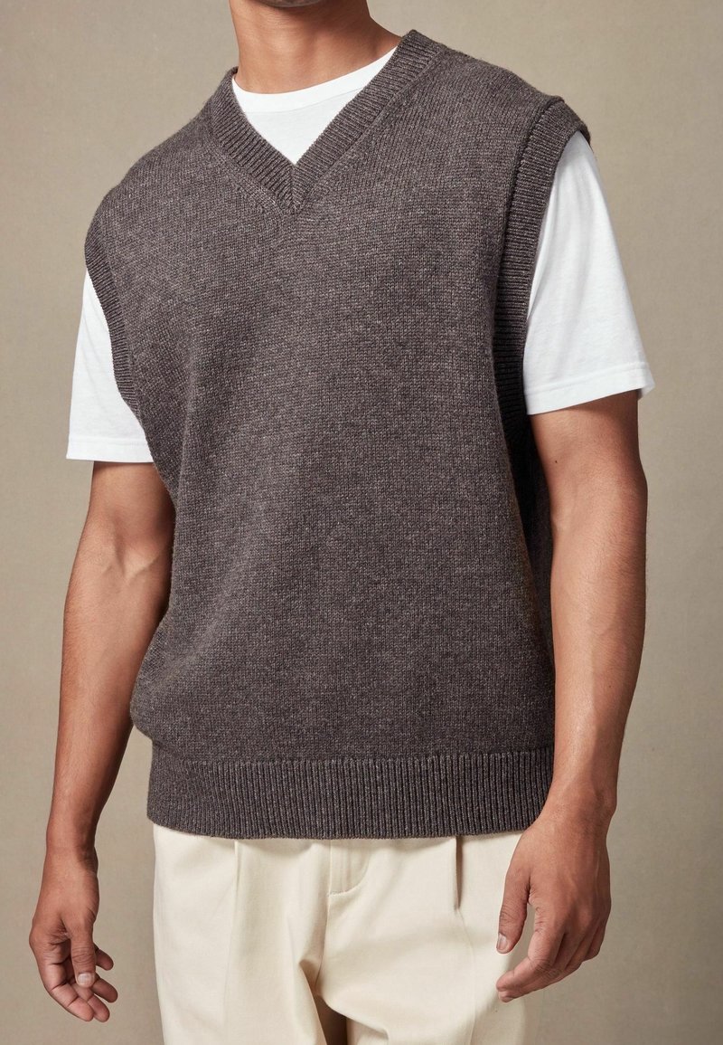 Gilet tricoté marron avec un décolleté en V, des manches courtes et un ourlet côtelé, porté par-dessus un t-shirt blanc, avec une texture douce.