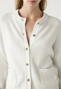 Valkoinen neulottu neuletakki, jossa on ribatut kaulukset, kultaiset napit ja etutaskut. Takissa on pehmeä pinta ja se on suunniteltu standardin mukaan.