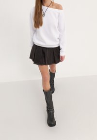 Sudadera blanca off-shoulder, falda de mezclilla oscura con pliegues y borde deshilachado, y botas negras hasta la rodilla con detalles de hebilla.