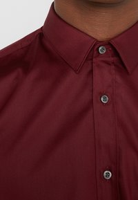 Camisa burdeos de botones, con una textura suave, cuello estructurado y botones a juego, que muestra un diseño ajustado con costuras precisas.