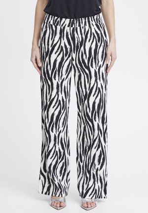 Kobieta ubrana w szerokie spodnie w czarno-białe paski zebry z elastycznym pasem oraz srebrne sandały na obcasie z odkrytymi palcami, stojąca na jednolitym białym tle.