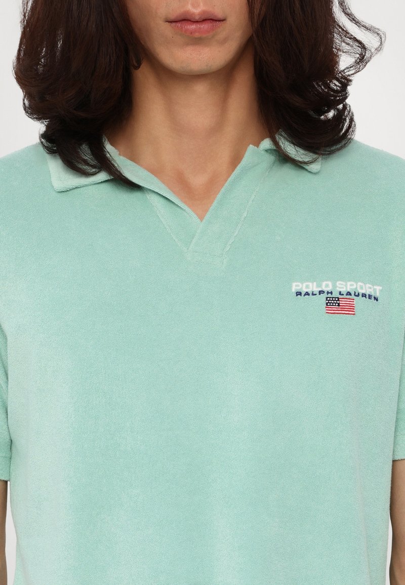Jasnozielona koszulka polo z krótkim rękawem, wykonana z miękkiego materiału frotte; ma kołnierzyk oraz haftowane logo w kolorze niebieskim i czerwonym.