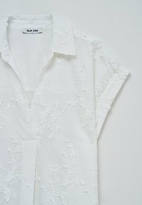 Camisa blanca de manga corta con bordado floral. Tejido suave, diseño con botones, presenta un cuello y mangas enrolladas, con costuras detalladas.
