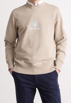 Mann trägt einen beigen Calvin Klein Pullover mit Logo und navyblauen Hosen, mit Händen in den Taschen, vor einem schlichten Hintergrund stehend.