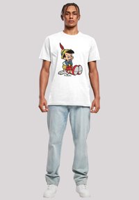 White cotton t-shirt featuring a cartoon Pinocchio graphic, with red, yellow, and blue details. Paired with light blue jeans and white sneakers.