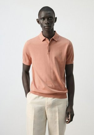 Hombre con una camisa polo de manga corta color salmón y pantalones beige claro, posando frente a un fondo blanco liso.