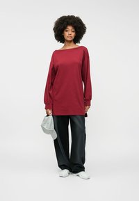 Μαρούλι oversized μπλούζα με μακριά μανίκια και στρογγυλή λαιμόκοψη, συνδυασμένη με μαύρο παντελόνι σε χαλαρή γραμμή. Το μοντέλο κρατά ένα ανοιχτό γκρι καπέλο.