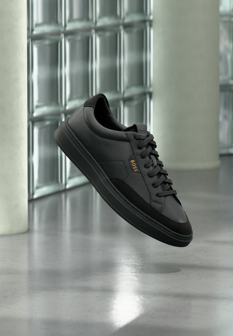 Sneaker basse en cuir noir avec lacets noirs et logo "BOSS" doré, affichée en suspension devant un mur en blocs de verre et un sol gris.