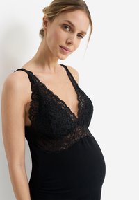 Μαύρο φόρεμα εγκυμοσύνης με μπούστο από δαντέλα με κυματιστές άκρες, βαθύ ντεκολτέ σε σχήμα V και εφαρμοστή γραμμή που προσαρμόζεται στην αυξανόμενη κοιλιά.