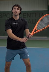 Tennisser in een zwart shirt en blauwe shorts, met een oranje tennisracket, voorbereid om te serveren op een blauwe binnenbaan.