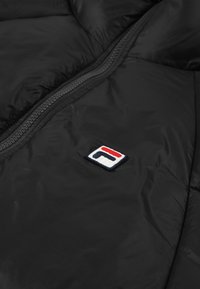 Svart puffjacka med central dragkedja och en liten broderad logotyp i rött, vitt och blått. Mjuk, slät textur.