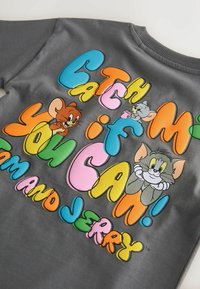 T-shirt gris avec texte coloré « Attrape-moi si tu peux ! Tom et Jerry » et personnages de dessin animé Tom et Jerry en poses ludiques.
