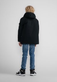 Svart vadderad jacka med huva, designad med en slät yta och en något längre bakdel. Buren med ljusblå jeans och svarta sneakers.