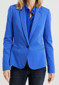 Blå kavaj i slät tyg, med en knapp, snedskurna slag och två framfickor, bärs över en blå mönstrad skjorta.