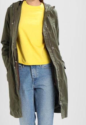 Olijfgroen gewatteerde parka met een ritssluiting aan de voorkant, met een zachte textuur, gecombineerd met een felgele t-shirt en lichtblauwe jeans.