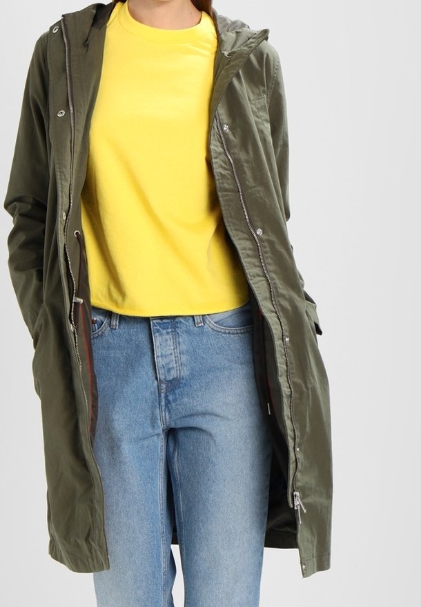 Olivově zelená parka s kapucí a zipem vpředu, která má měkkou texturu, doplněná jasně žlutým tričkem a světle modrými džíny.