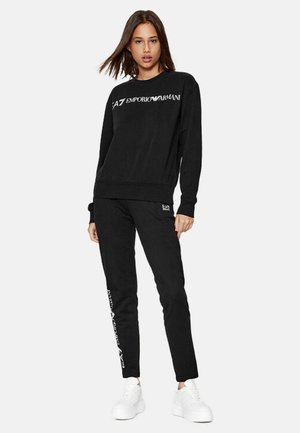 Felpa nera con scollatura rotonda e logo bianco. Abbinata a pantaloni della tuta neri con dettagli del logo sulla gamba. Sneakers bianche.