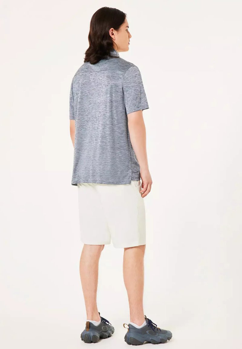 Persona joven con cabello de longitud media que lleva una camiseta gris de manga corta, pantalones cortos blancos y zapatillas grises, de pie de perfil.