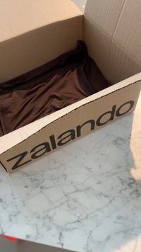 Papptförpackning från Zalando med en stor svart logotyp, delvis öppen för att avslöja krumlat brunt tyg inuti. Ligger på en marmorytan.