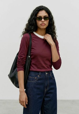 Donna con capelli lunghi e ricci che indossa occhiali da sole, una camicia bordeaux a maniche lunghe, jeans scuri e porta una borsa a tracolla nera.