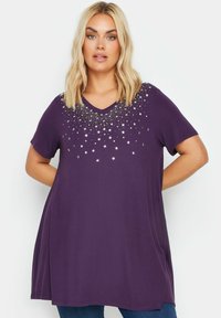 V-kauluksellinen violetti t-paita, lyhyet hihat ja tähkäkuvioinen helmikoristelu rinnassa. Pehmeä kangas ja rento istuvuus.