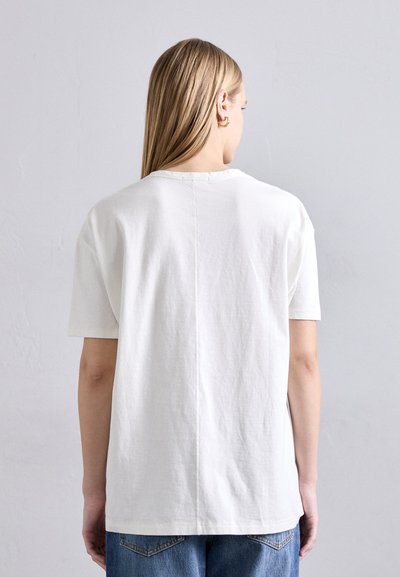 White cotton t-shirt with short sleeves, crew neckline, and a straight cut. Features a central seam and soft texture. Worn with blue jeans.