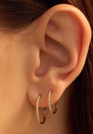 Gouden creolen met een gedraaide ontwerpen, hangend aan het oorlel van een model, met een glanzende afwerking en ronde vorm.