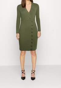 Femme portant une robe en maille côtelée vert olive à manches longues, col en V profond, détails de boutons sur le côté, et escarpins noirs à brides sur fond blanc.