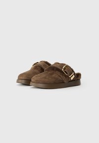 Pantofi de casă din suede maro, slip-on, cu căptușeală din blană, cataramă aurie ajustabilă în partea de sus și talpă din cauciuc plat, prezentati pe un fundal simplu deschis.